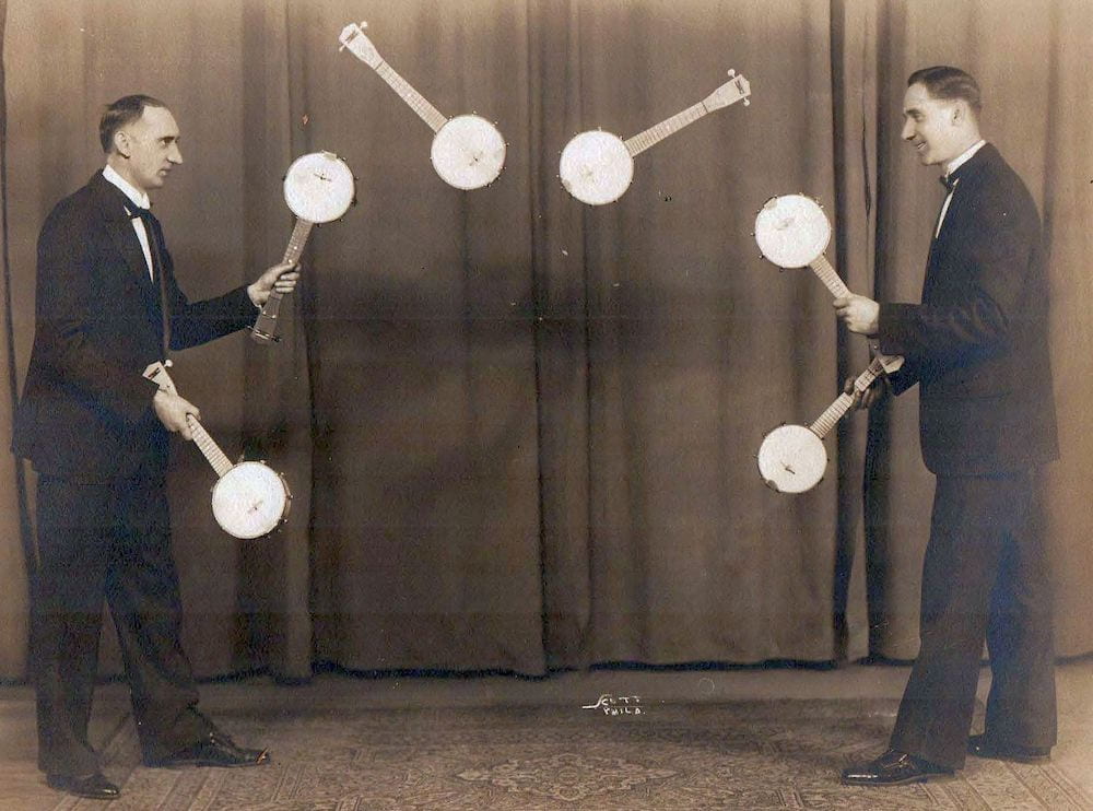 A photograph of two men juggling 6 banjos between them.