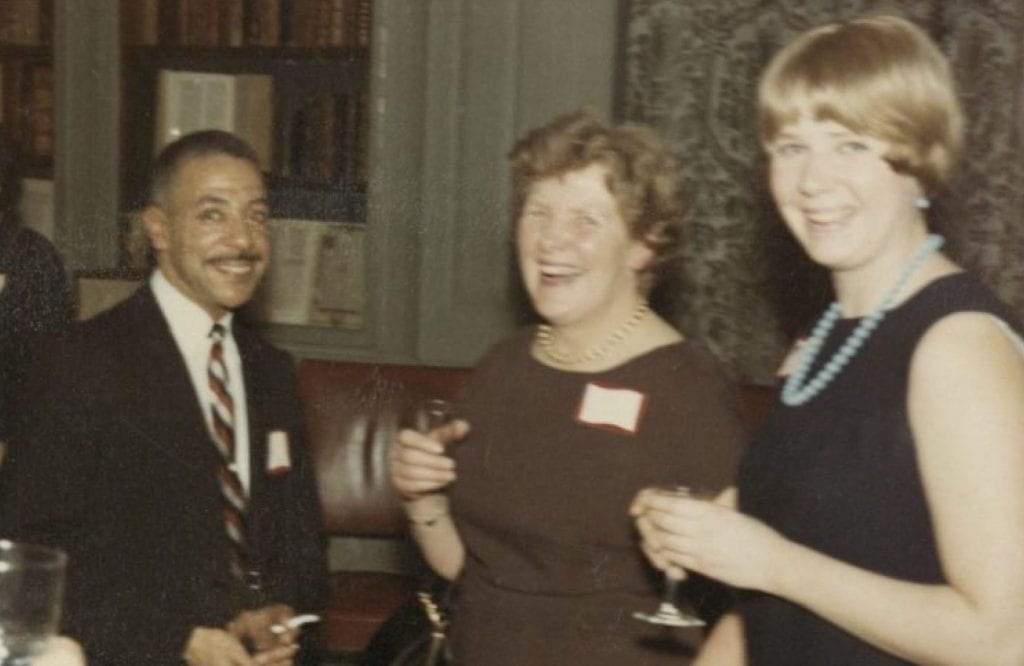 A Black man in a suit holding a cigarette and two white women holding drinks in a party setting.