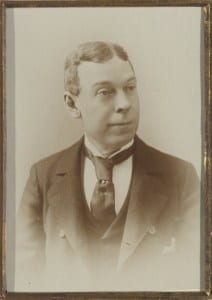 A man in suit, waistcoat, and black tie, clean-shaven with his hair parted in the middle.