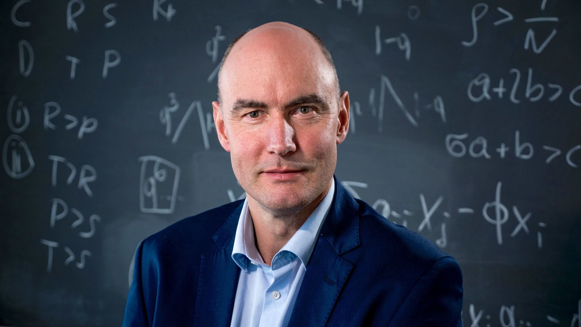 A headshot of Harvard math and biology professor Martin Nowak standing in front of a blackboard covered with mathematical formulas.