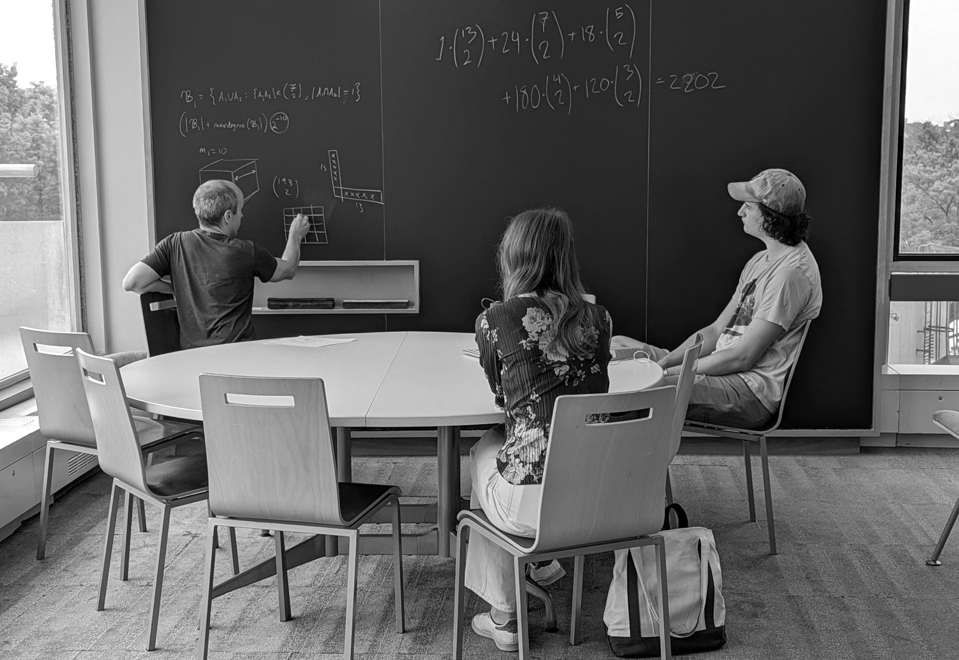 A black and white photo of a math instructor writing formulae on a blackboard in front of two students sitting at a round table.