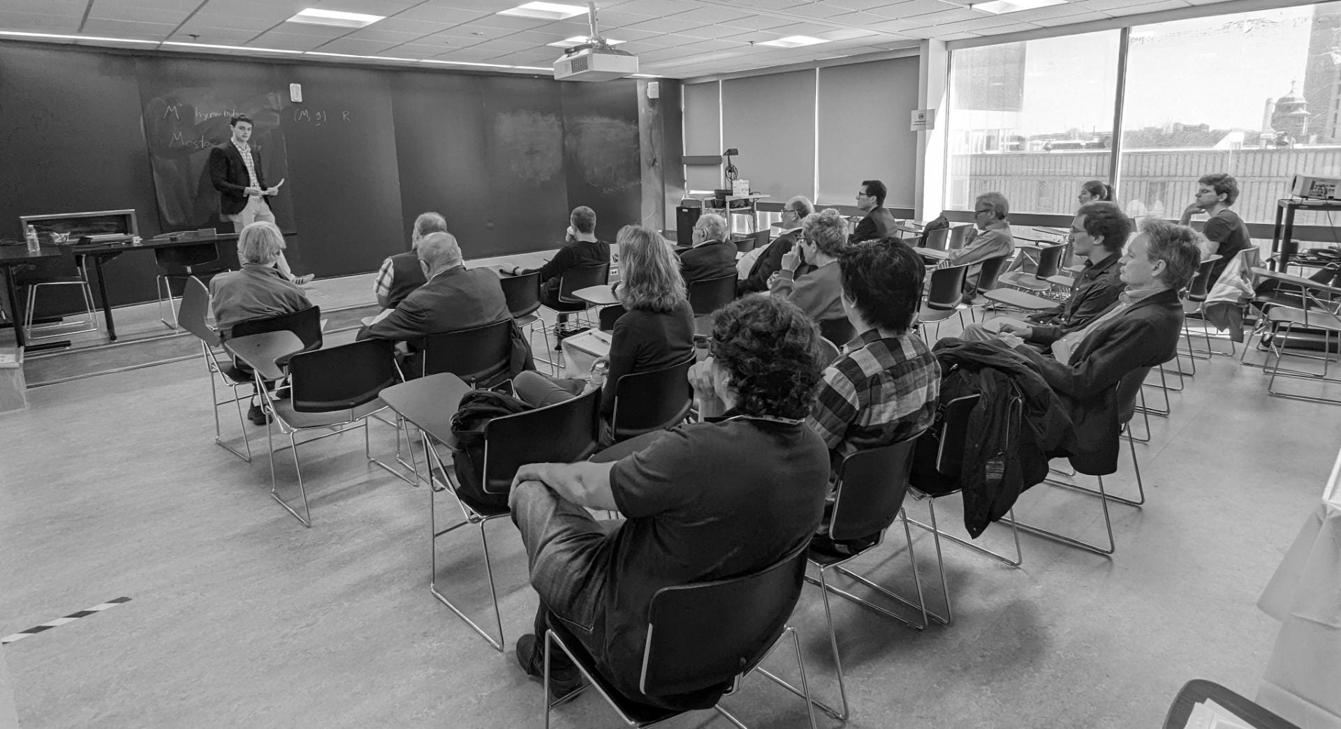 A black and white photo of a student giving a mathematical presentation in a classroom in front of faculty members.